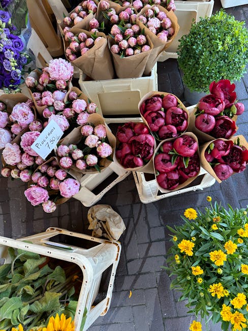 An outdoor display of fresh flower arrangements and potted plants, including multiple bouquets of pink peonies wrapped in brown paper, arranged in white plastic crates on a brick pavement. Some bouque