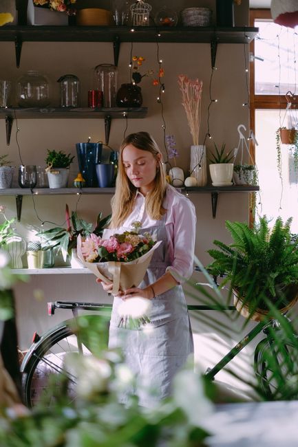 A young woman with light brown hair, dressed in a white blouse and apron, stands indoors in a well-lit flower shop or studio, holding a bouquet of fresh flowers wrapped in brown paper. The bouquet fea