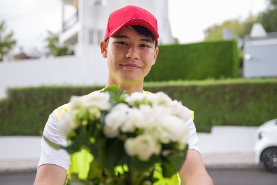 A young delivery person wearing a red cap and high-visibility vest is smiling while holding a bouquet of fresh white flowers towards the camera. The bouquet features a variety of light-colored blooms 