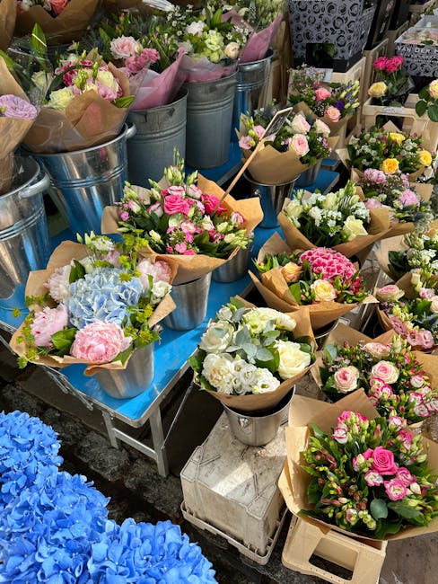 An assortment of fresh floral bouquets and arrangements displayed on a table, featuring roses, hydrangeas, and other blossoms in shades of pink, white, and blue. Some flowers are wrapped in brown craf